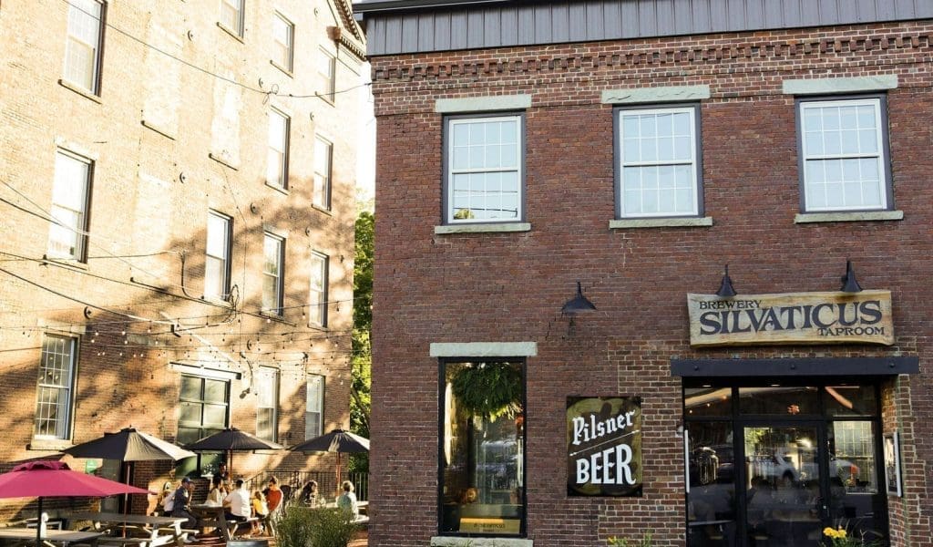 Frontal view of a red brick brewery building named Silvaticus Taproom with a Pilsner Beer sign in the front on a sunny day in Amesbury MA with people sitting outside to the left under tables with umbrellas