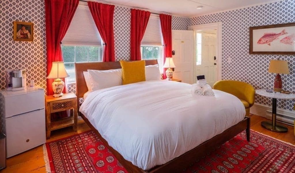 A room with blue & white printed wallpaper with framed pictures on the wall, bright red open curtains on each window, 2 wooden nightstands with an antique looking lamp on each, a mini fridge, a yellow chair, a bed with white bedding and one yellow pillow with clean towels placed upon it, and a printed red carpet underneath the bed at Boutique Inn called the Sally Webster Collection in Rockport MA