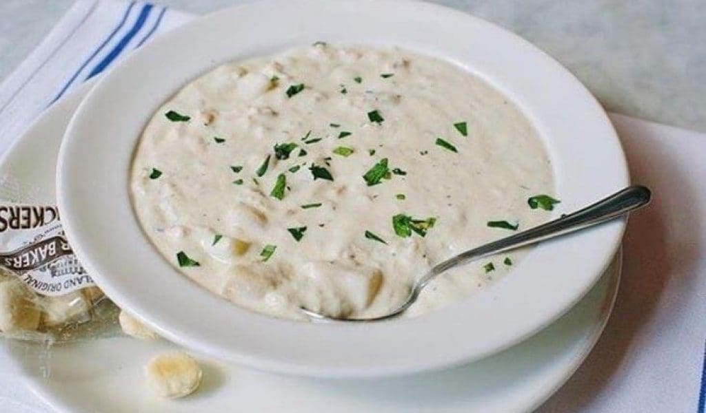 Freshly prepared New England Clam Chowder from Turner's Seafood in Salem, MA in a white bowl with a silver spoon in the chowder and oyster crackers on the saucer.